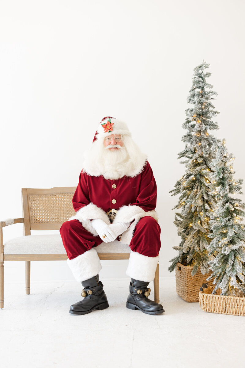 Pictures with Santa in Tulsa with photographer, Caroline Eidson Portraits. Clean white studio with Santa on a bench and Christmas tree. 