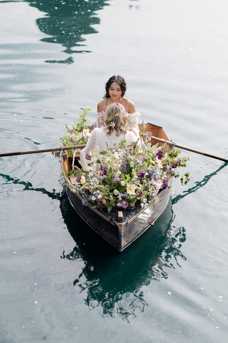 Two brides in a boat filled with pretty flowers  in the water at Euridge manor wedding venue in  the Cotwolds