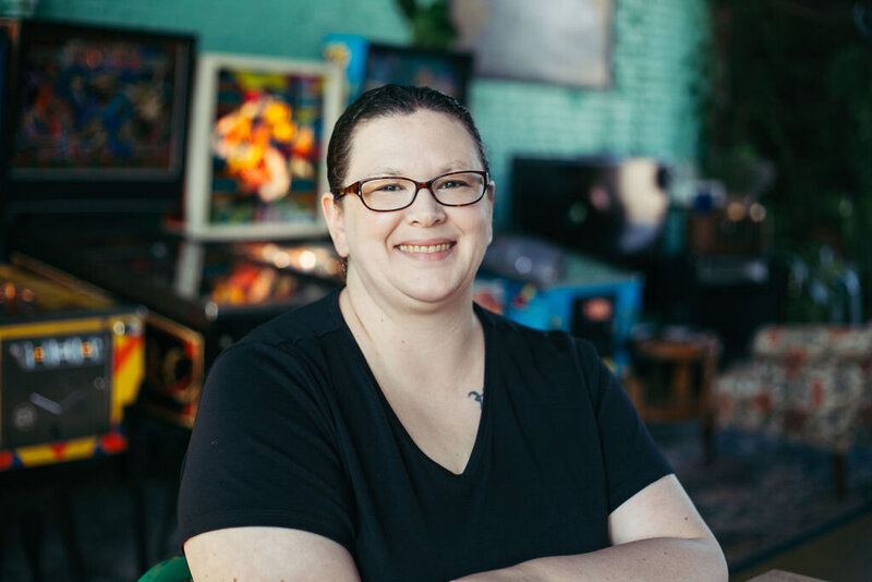Kristen, owner of Clockwork Scribe Consulting, smiling at camera with pinball machines in background.
