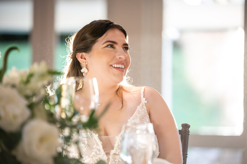curvy white bride in a spaghetti-strap gown sitting down at a sweetheart table with white flowers on top staring and smiling off camera