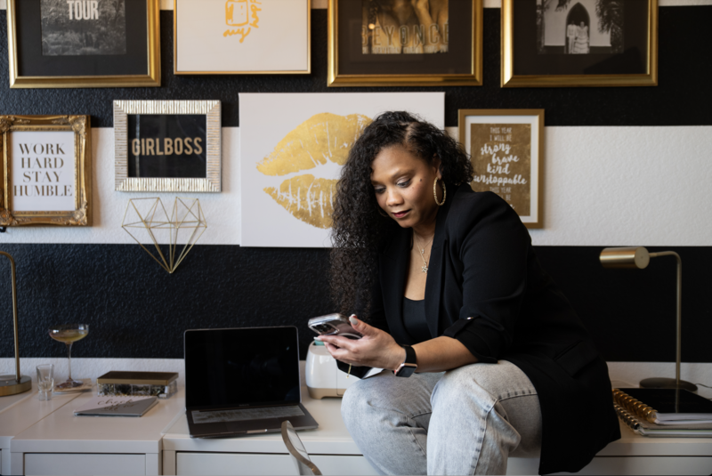 candice wearing a black blazer and jeasn, sitting on a desk in her office and looking down at her phone.