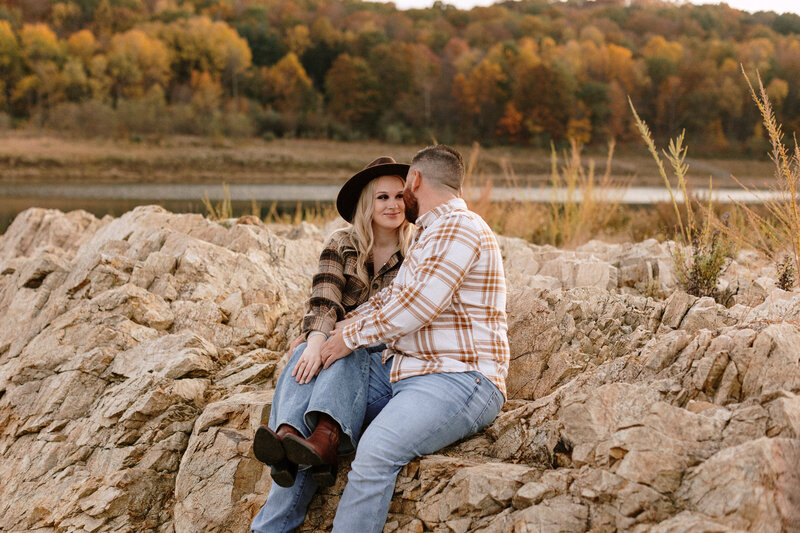 A couple embraces by a serene lake, surrounded by vibrant fall foliage, capturing the warmth of their engagement.