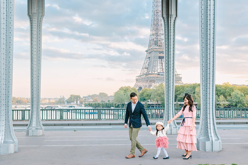 Family of three — mother, father, and daughter — holding hands while walking near the Eiffel Tower at Bir-Hakeim during a Paris family photoshoot.