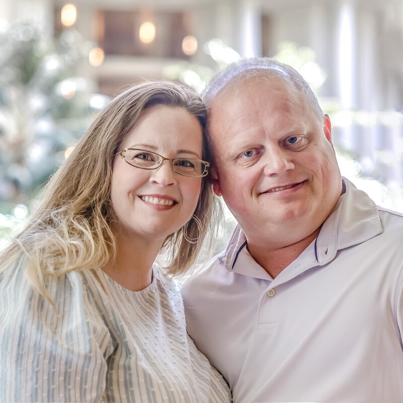 A smiling husband and wife team from North Carolina pose closely together, embodying warmth and connection. The woman, with long wavy hair and glasses, wears a light top, while the man, dressed in a light collared shirt, stands beside her with a friendly expression. This image captures the essence of love and partnership, perfect for showcasing the intimate moments often highlighted in North Carolina wedding photography. The softly lit background features blurred greenery, enhancing the couple's joyful demeanor.