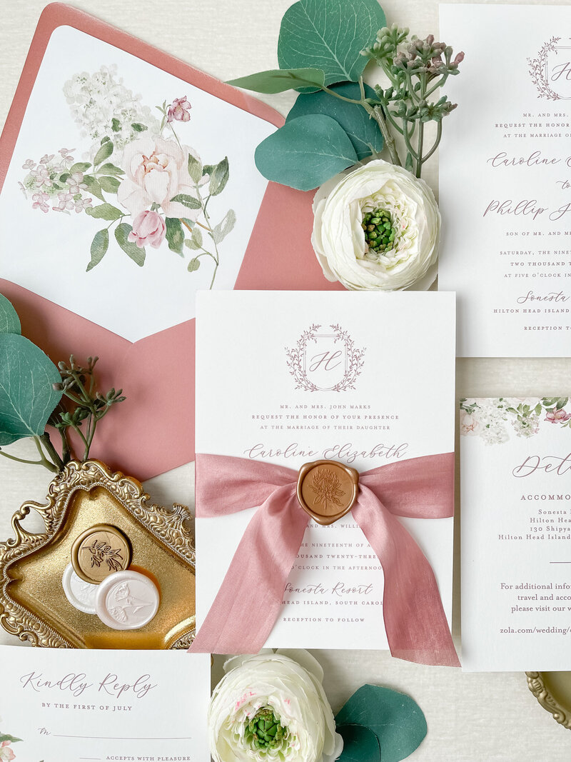 Caroline and Phillip's custom wedding invitation suite that includes a pink crest with monogram H, a pink silk ribbon with gold wax seal, and a pink envelope with floral watercolor on envelope liner