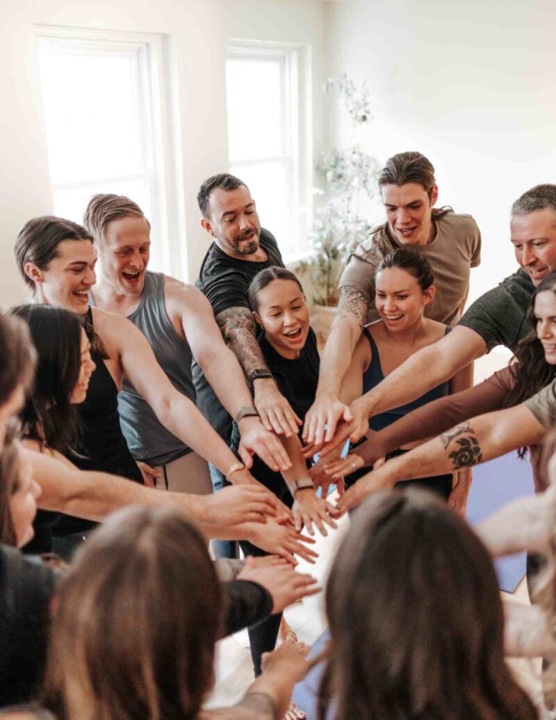 A diverse group of people in a breathwork class standing in a circle, placing their hands together in a show of unity and shared energy.