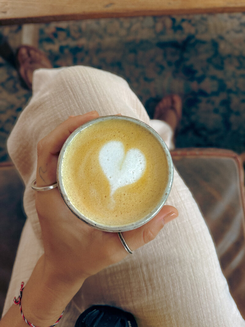 woman wearing beige pants with her legs crossed holding a cup of coffee with a heart in the foam 