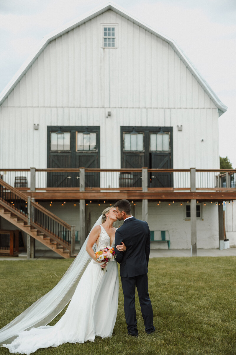 Bride and groom kiss in front of white barn at Lincoln Farmstead