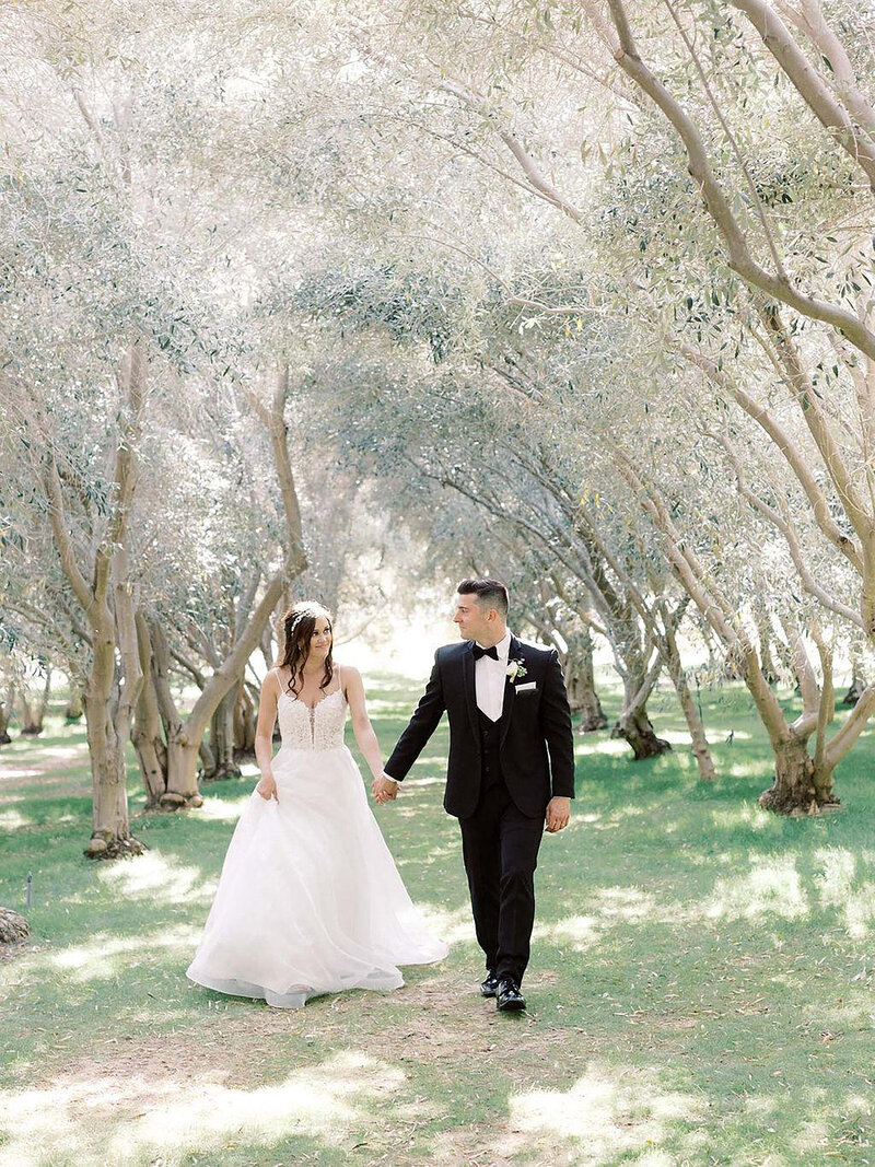 6-radiant-love-events-Temcula-bride-groom-holding-hands-looking-at-eachother-walking-in-beautiful-tree-orchard-updated-romantic-elegant-timeless