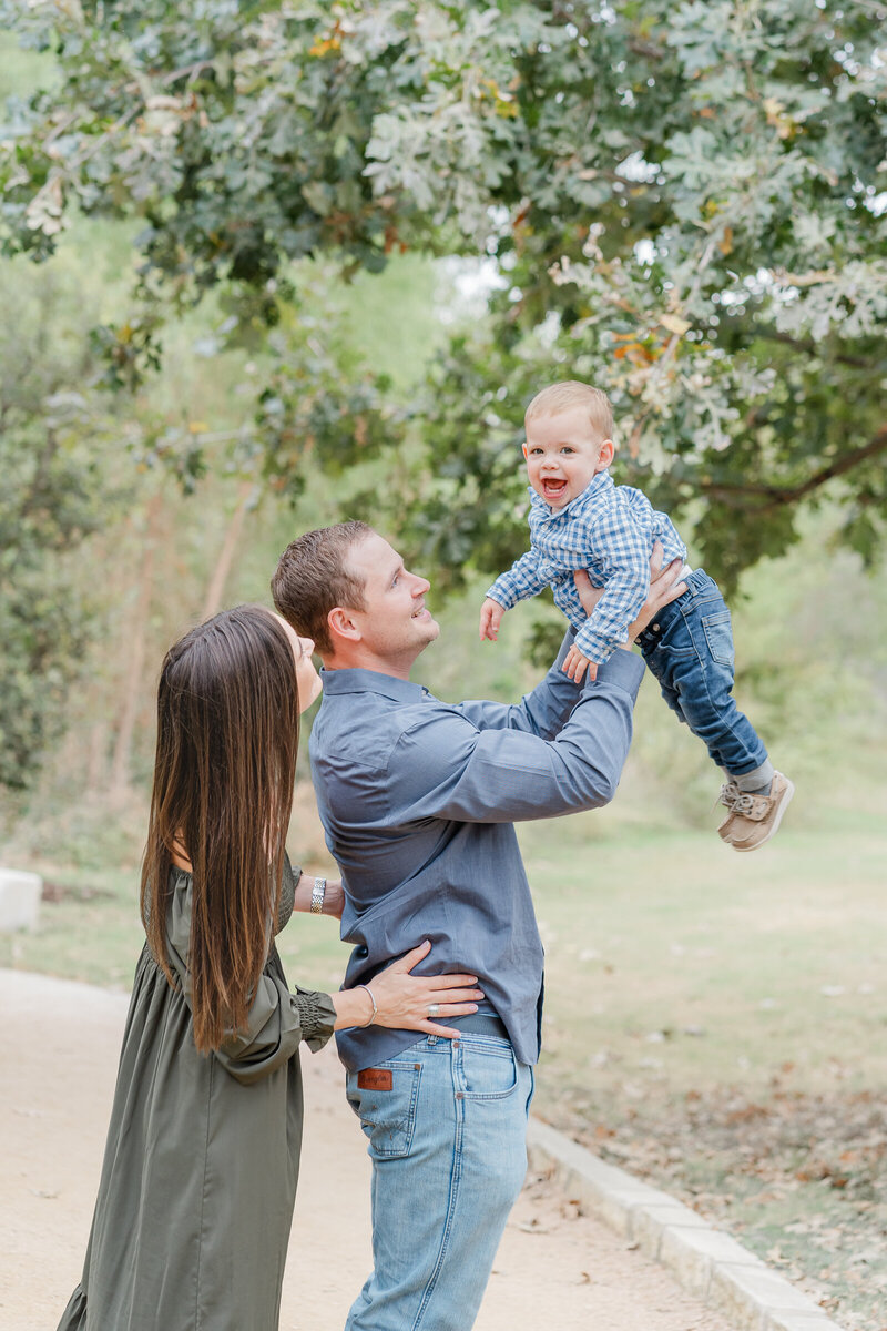 Austin, TX Family & Newborn Photography | A mom and dad stand on a path at Katherine Fleischer Park. The dad holds their baby boy up in the air while his parents look up at him and smile.