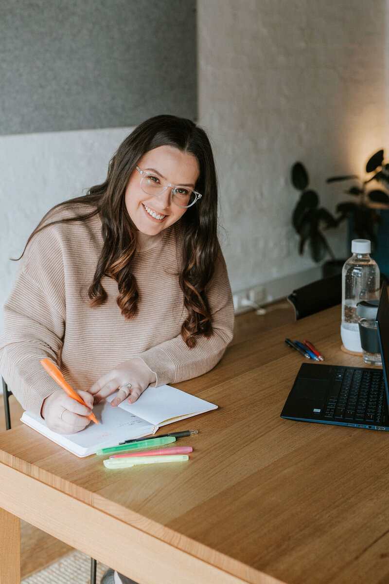 Kirsten from KP Virtual Assistance smiling warmly at her desk, highlighting a professional yet approachable demeanor. She is writing in a notebook surrounded by highlighters, pens, and a laptop, representing her organised and creative approach to helping photographers streamline their businesses.