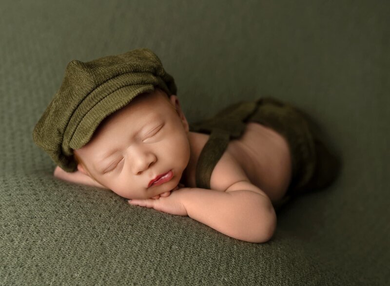 newborn boy wearing green overalls and green courdoroy pageboy hat posing for newborn photoshoot