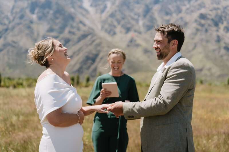 Bride and groom exchanging rings and smiling joyfully during an outdoor wedding ceremony, with a celebrant officiating in front of a backdrop of majestic mountains. Melissa Clark Photography