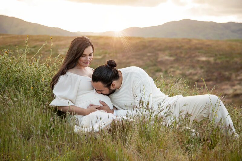 Couple sitting in field, expecting father kissing her belly | California Maternity Session