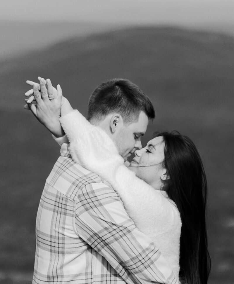 A black and white image of a couple embracing closely against a blurred mountainous background captures the tender moment with the skill of a New England Wedding Photographer. The woman, in a fluffy sweater, wraps her arms around the man's neck as they gently touch foreheads, eyes closed and smiling softly.