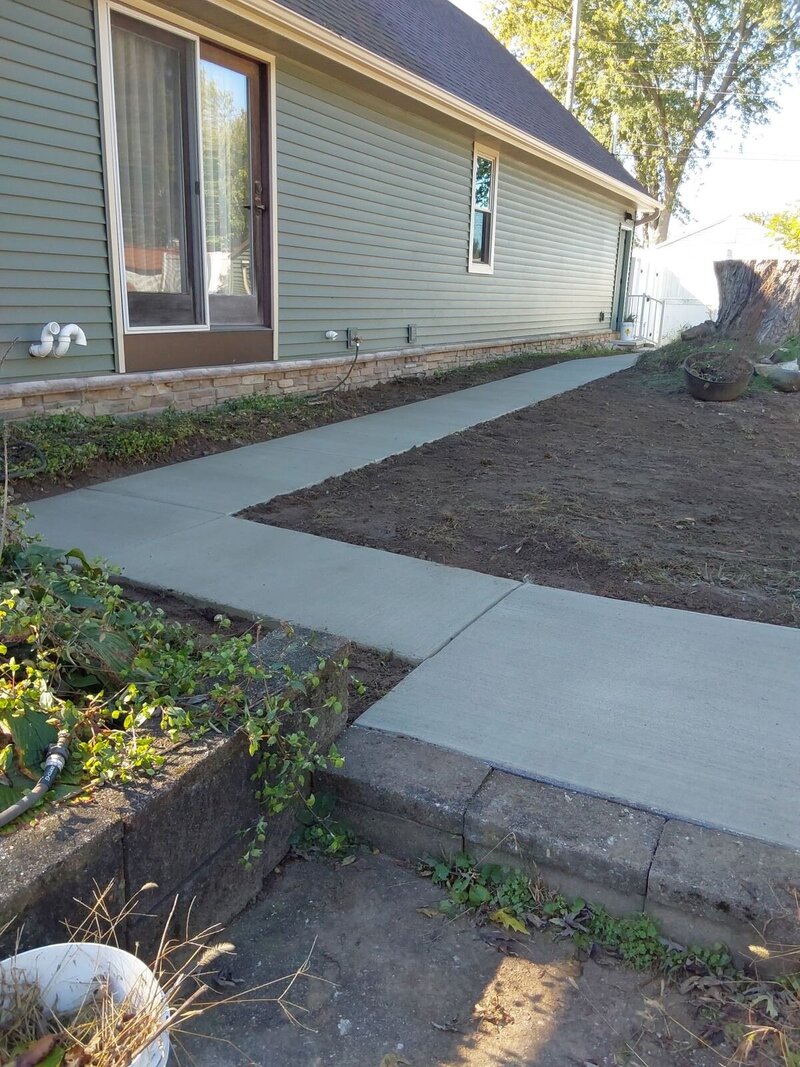 New concrete walkway wrapping along the back of a house with new siding, stepping down from a block retaining wall in Osceola, IN