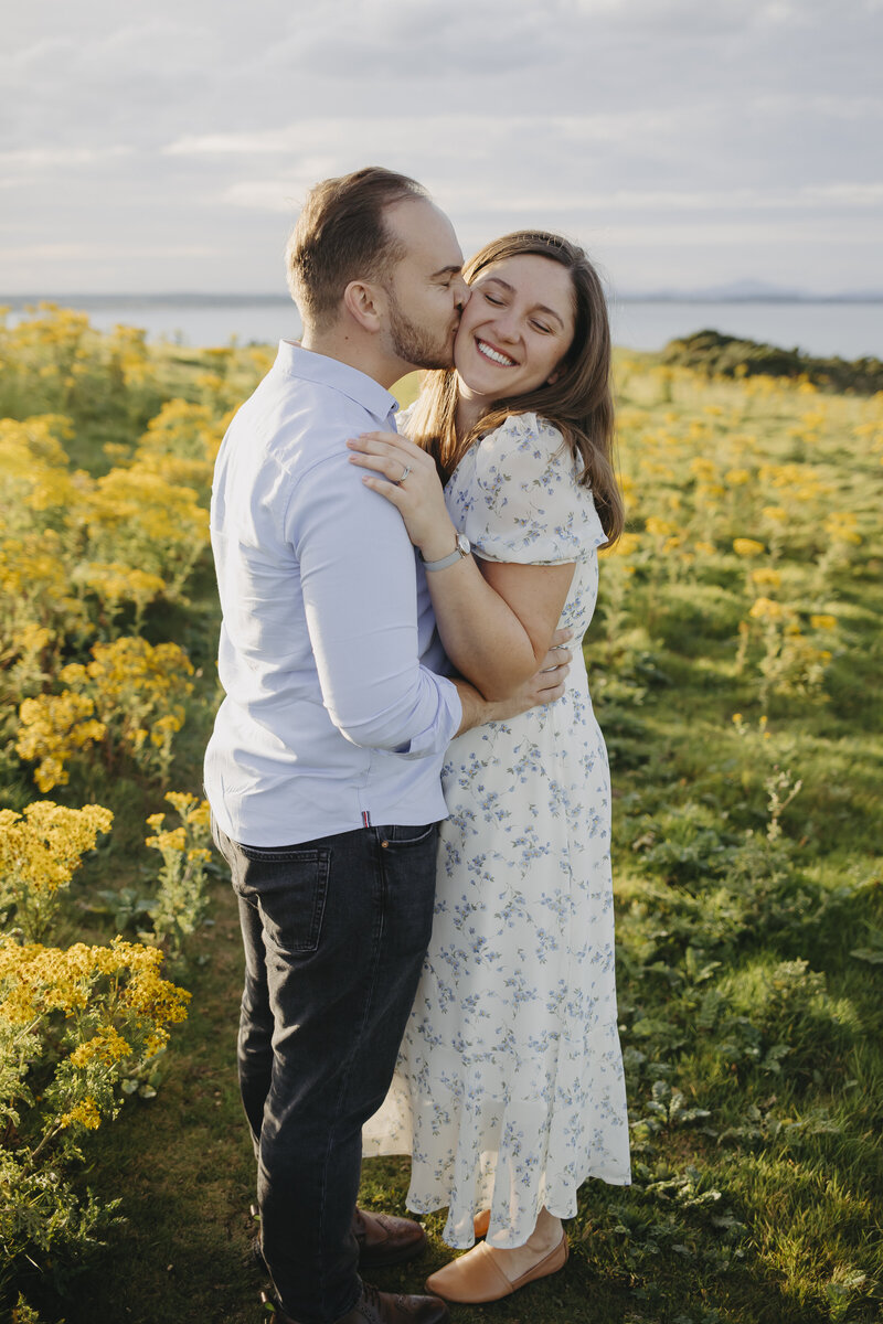 Couple photoshoot on the cliffs of Clogherhead, with the man kissing his partner on the cheek amid a field of yellow wildflowers.
