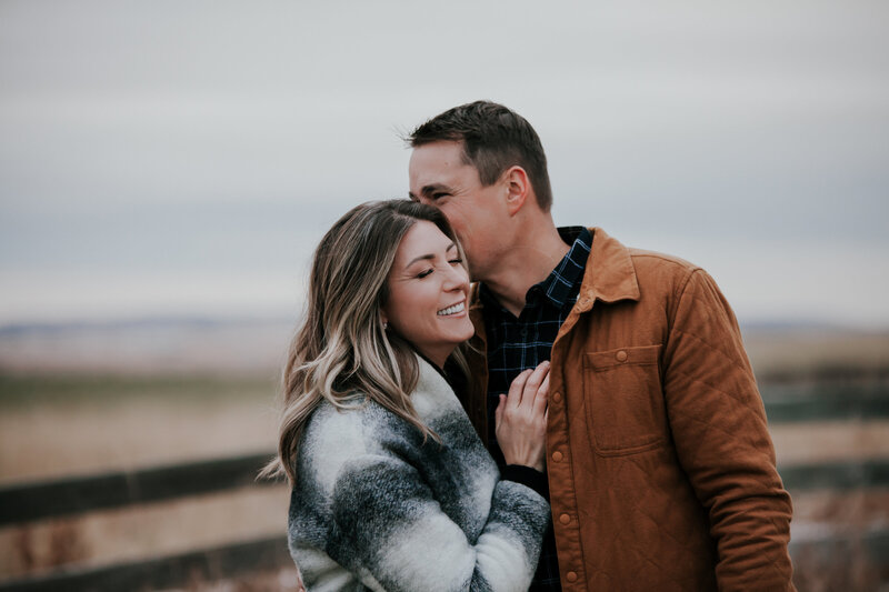 A man kisses his fiance on the cheek during their enagagement session in a Alberta foothills field. She has her eyes closed and is smiling with her hand on his chest