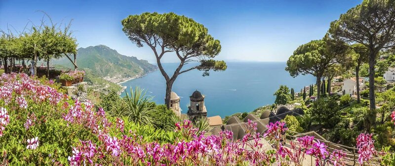 Scenic coastal view of the Amalfi Coast with pink flowers, tall trees, and blue sea under a clear sky.