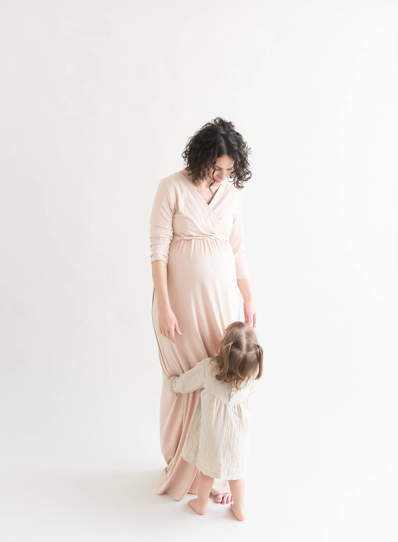 pregnant mom wearing a client closet light beige dress looking down at toddler hugging her leg in studio setting