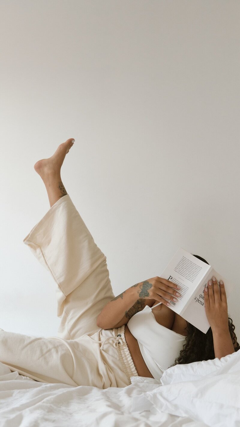 Person lying on a bed reading a book, with one leg playfully raised. They're wearing beige pants and a white top, creating a relaxed atmosphere.