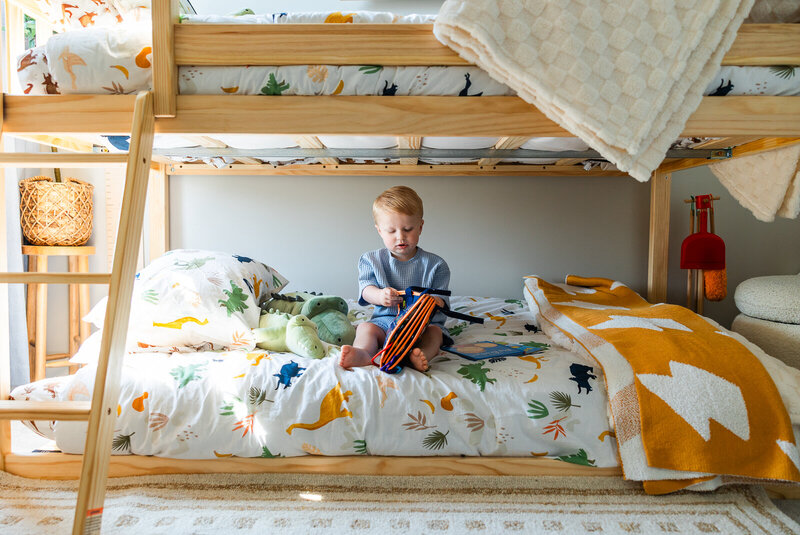 Toddler boy sitting on his bed looking through books with the sunlight coming through the window. 