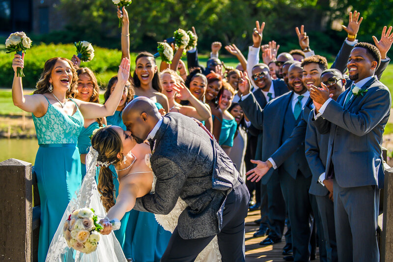 A joyful wedding scene with a bride and groom kissing passionately while surrounded by their cheerful wedding party. The bride, in a lace gown with a flowing veil, and the groom, in a dark patterned suit with a black bow tie, are at the forefront. The bridesmaids wear teal dresses and the groomsmen are in dark suits with green ties. Everyone is smiling broadly and raising their arms in celebration, with a lush green park background.