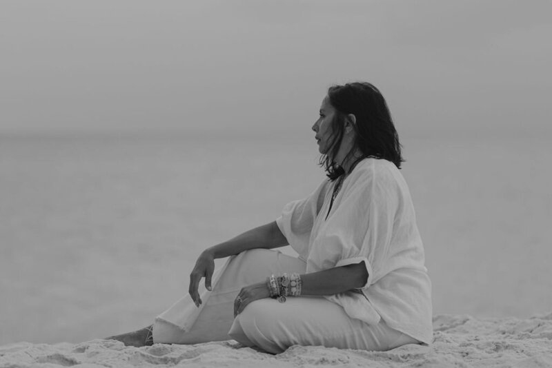 Side view of woman sitting on beach in white pants and white shirt wearing lots of bracelets with shoulder length very dark brown hair looking off at the beach