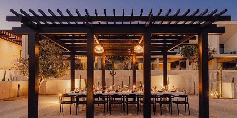 An elegant outdoor dining setup at Nobu Los Cabos, featuring a candlelit table under a modern pergola with warm lighting and desert landscaping. 