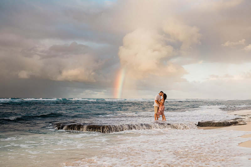 couple standing on a rock kissing in front of rainbow at the beach