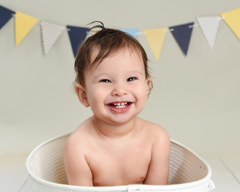 A smiling baby sitting in a white tub with colorful yellow, blue, and navy bunting in the background.
