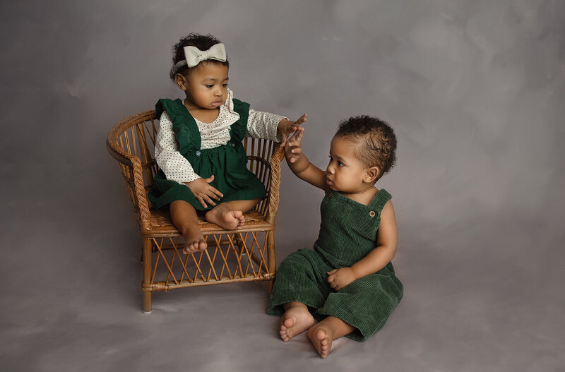 Twin brother and sister in green dress and overalls sit and touch hands in a studio floor