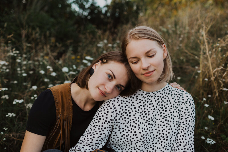 Aikuiset siskot istuvat maassa vierekkäin lähellä toisiaan ja nojaavat päitä yhteen Suomenlinnassa Helsingissä