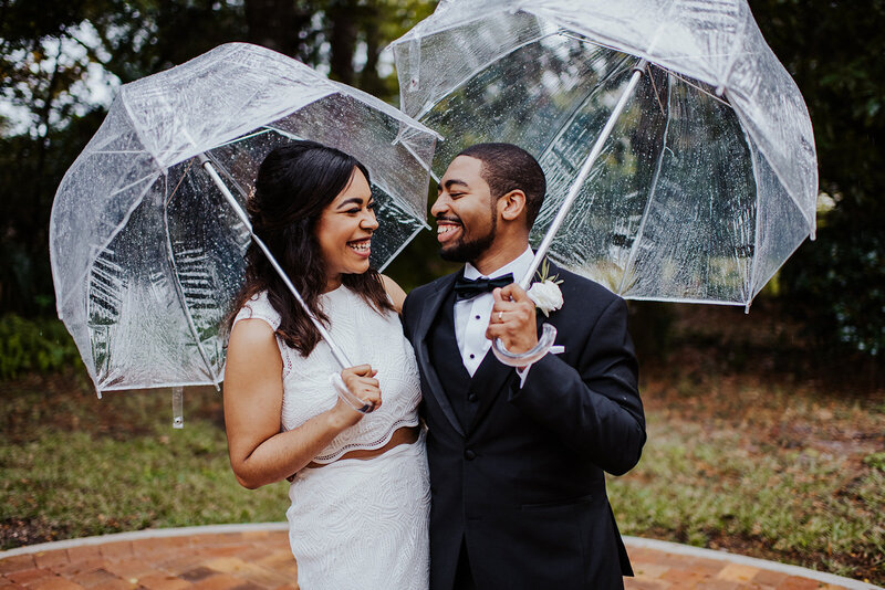 Bride sports a chic two piece set smiles at groom in a tux as they stand outdoors under clear umbrellas.