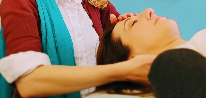 A colorfully dressed Reiki Master has their hands on a clients forehead and shoulder to deliver energy healing. 
