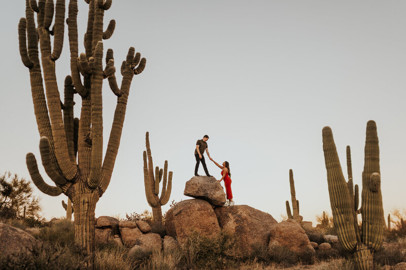 A couple stands on large boulders in the desert, surrounded by towering saguaro cacti, with one person helping the other balance atop the rocks.