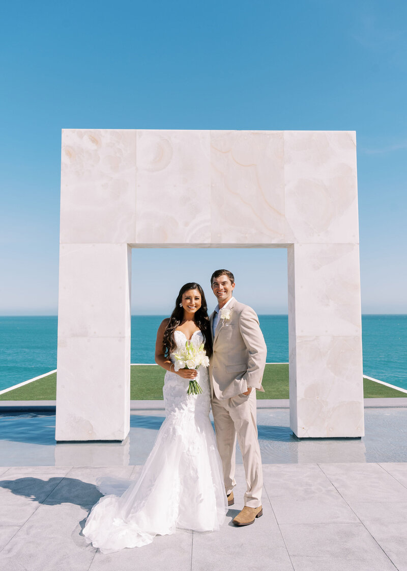 bride and groom standing in front of an archway with the ocean in the background