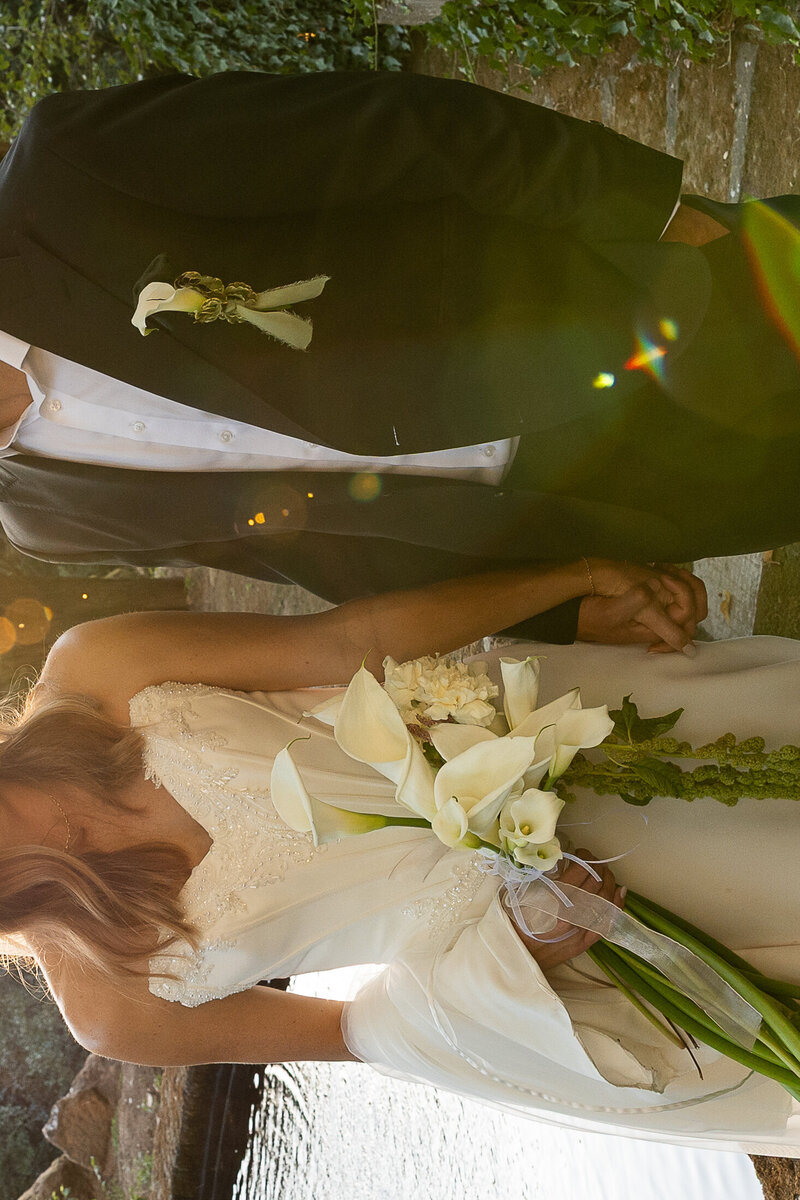 Film-style photo of a couple standing close together, bride is gently holding a bouquet of white calla lilies, their faces softly out of focus.