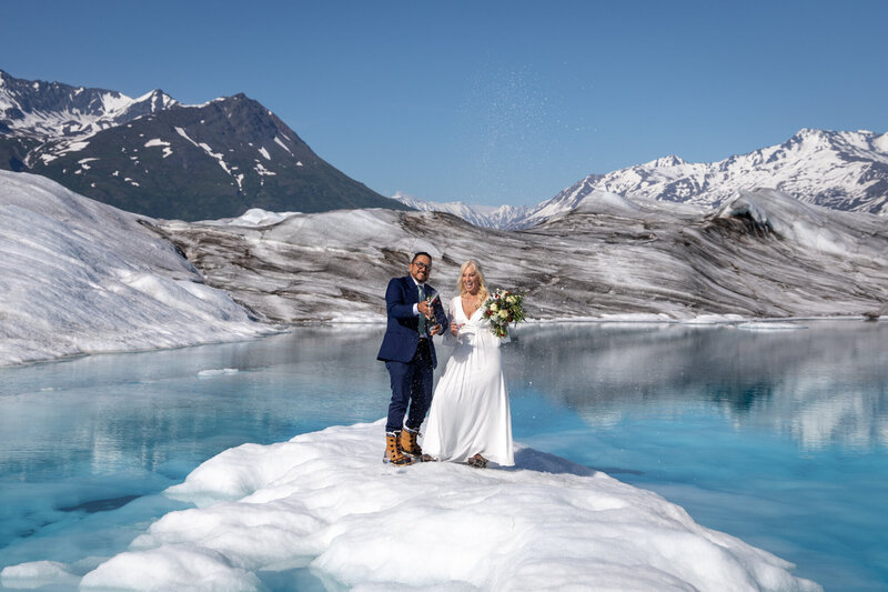 Bride and groom stand next to a bright blue glacial pool, the helicopter waits while they pop champagne and toast.