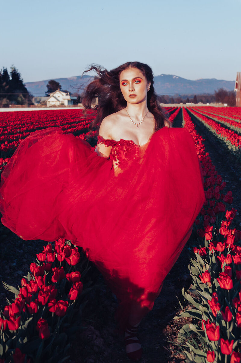 Woman in off-the-shoulder red tulle gown runs through field of red tulips.