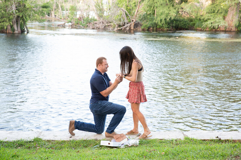 An Austin wedding photographer captures the intimate moment when a man kneels down next to a lake and proposes to a woman.