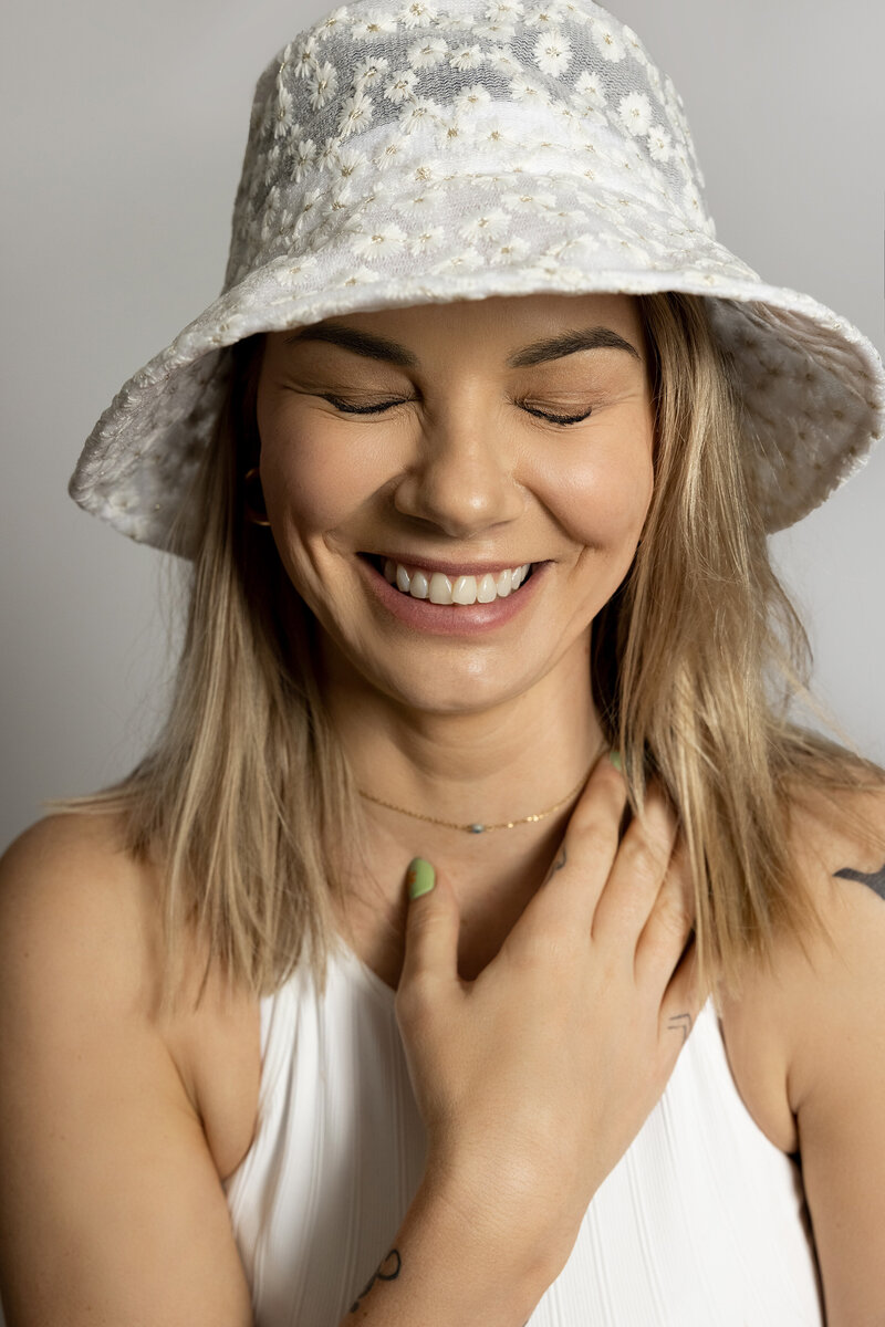 Best Headshot Photographer in London, Ontario. Woman in bucket hat is laughing at the camera with her eyes closed. She has one hand resting on her collarbone.