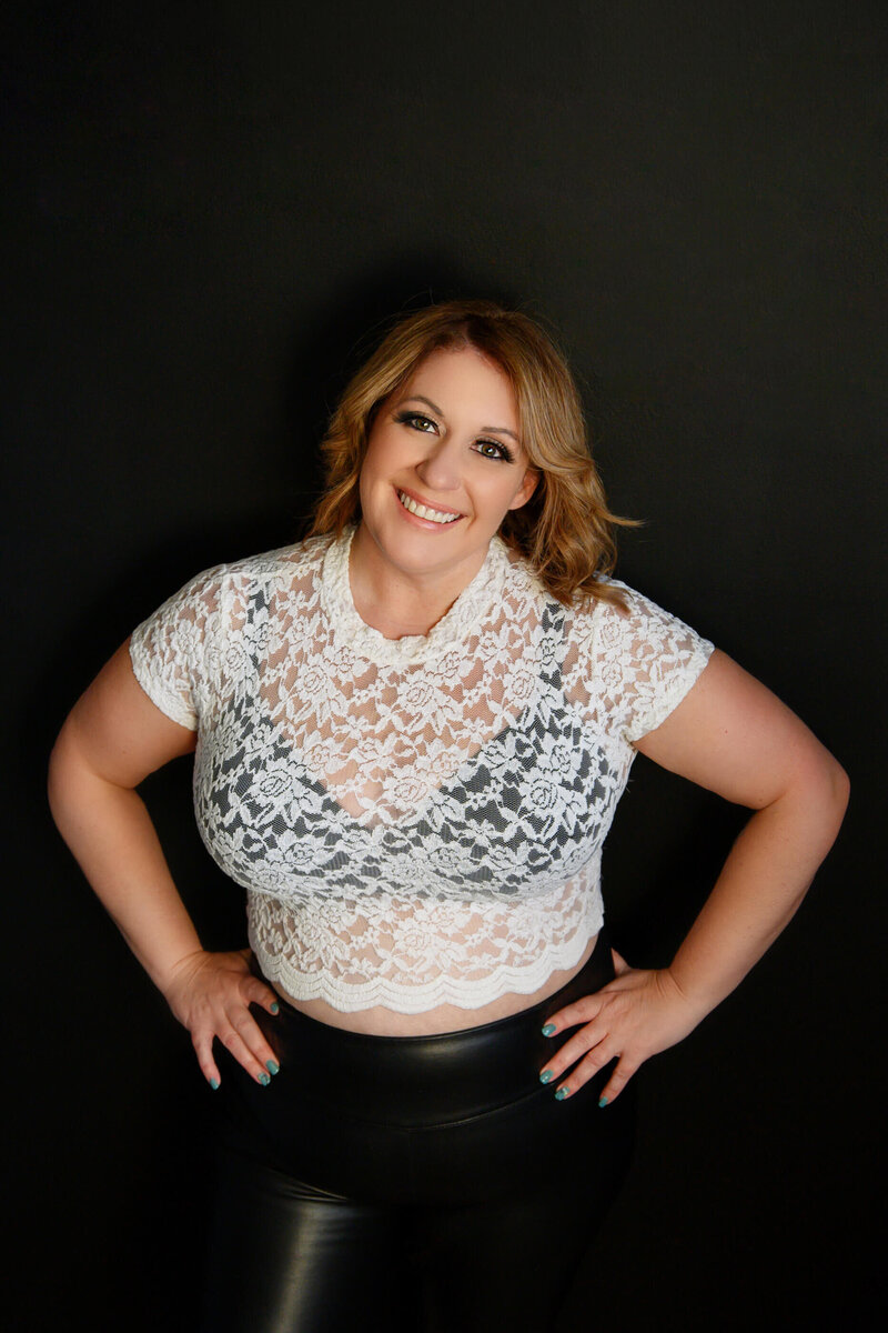 Amanda Gibson Owner of Gibson Boudoir wearing a white lace crop top leaning against a black wall with her hands on her hips.