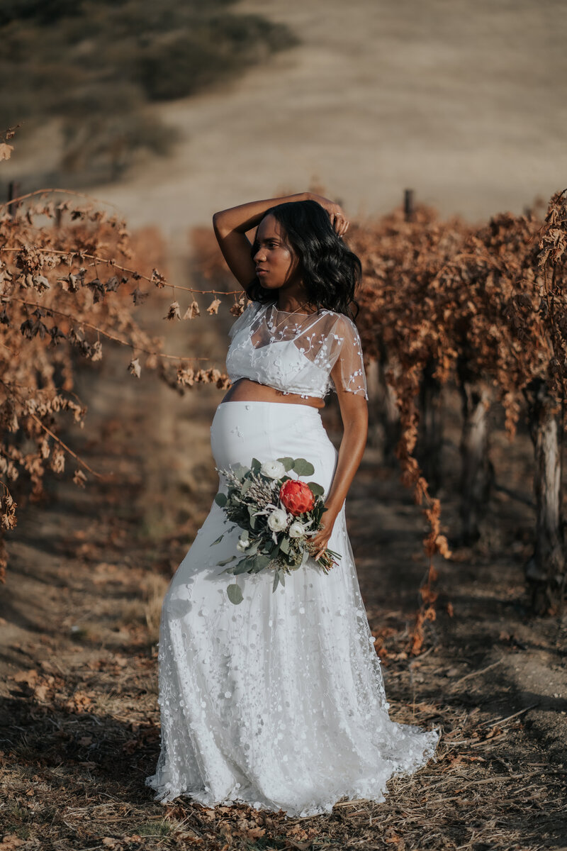 A pregnant bride in a wedding dress, holding a bouquet and standing in a row of the Cass Winery vineyard