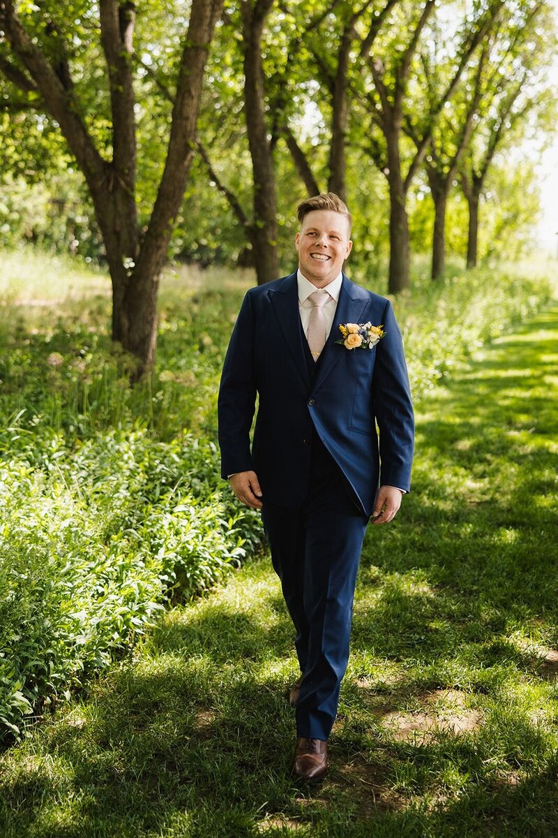 Groom in blue suit walking up a aisle surrounded by trees.