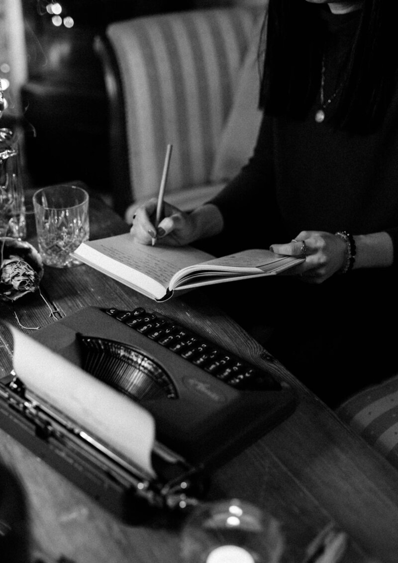 Black and white image of a woman writing in a journal beside a typewriter