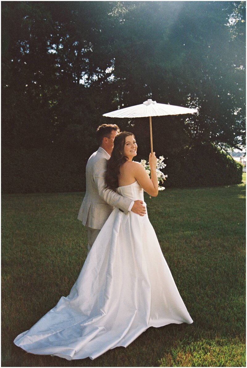 A moment captured by a Wilmington wedding photographer of a bride walking with her groom's arm around her waist in a garden holding a white umbrella