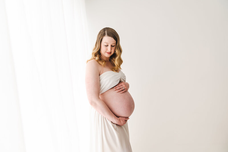 A radiant expecting mother in a white dress tenderly cradling her baby bump against a bright, airy backdrop.