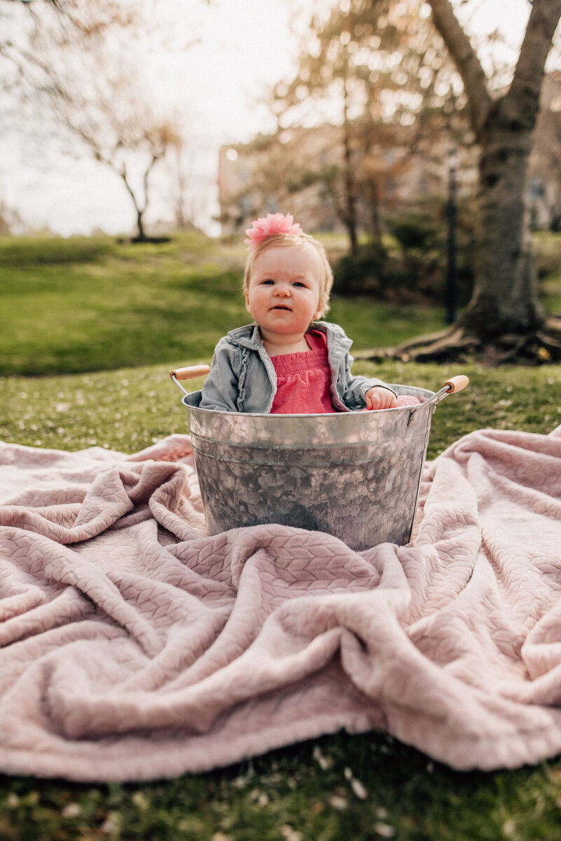 Baby sits in a big bucket on a blanket outside.