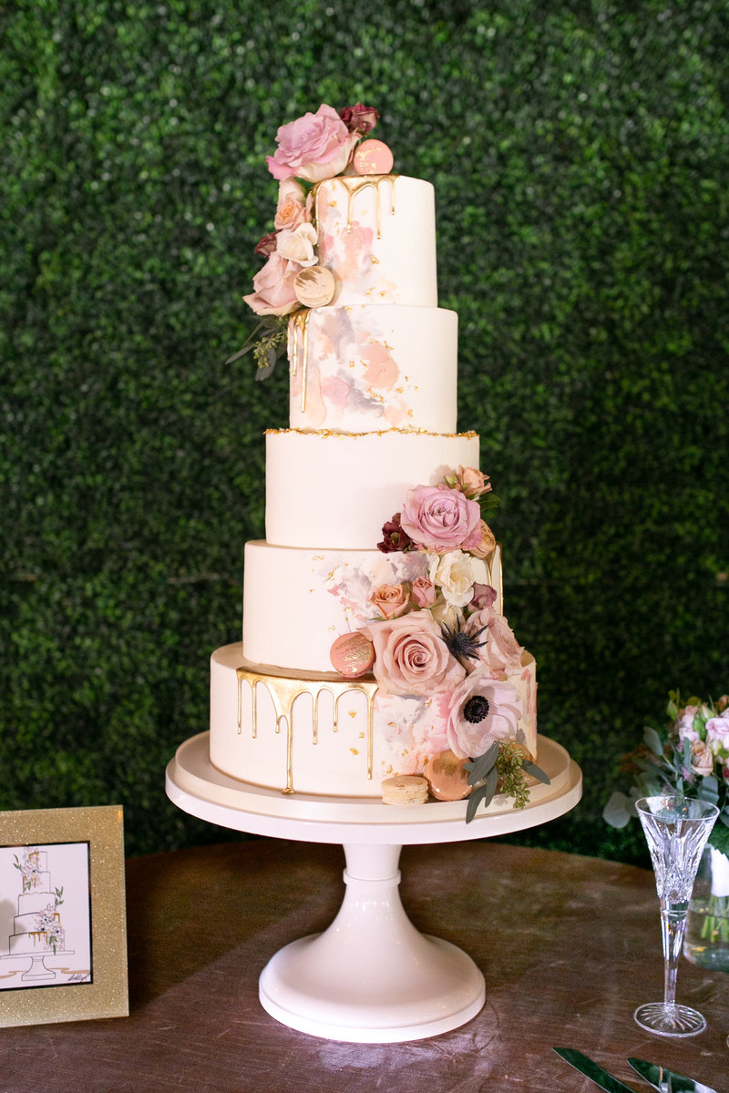 Modern white buttercream wedding cake with pastel palette knife icing gold chocolate drips deckle edge macarons and colorful flowers.